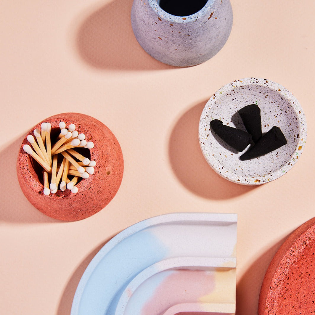 A coral terrazzo matchstick holder next to a white terrazzo incense holder.