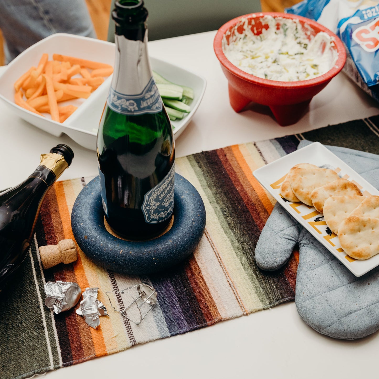 Cobalt blue terrazzo wine bottle coaster, styled on a table with a few sparkling wine bottles, the oven mitt trivet, and food.